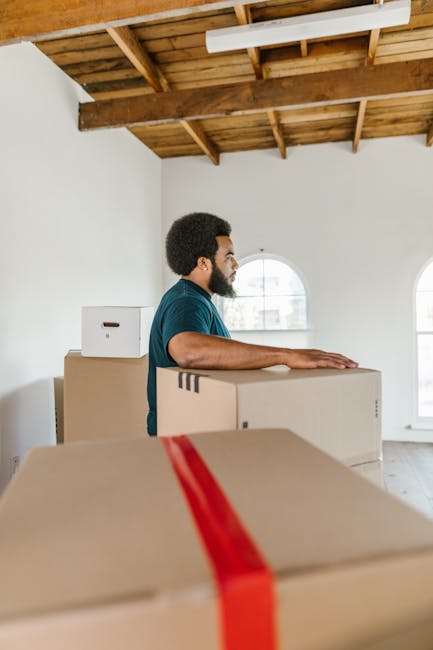 A young woman with long brown hair smiling at the camera while holding an open cardboard box labeled 'GLASS' in red, indicating fragile contents, during a home relocation process. She is standing indo